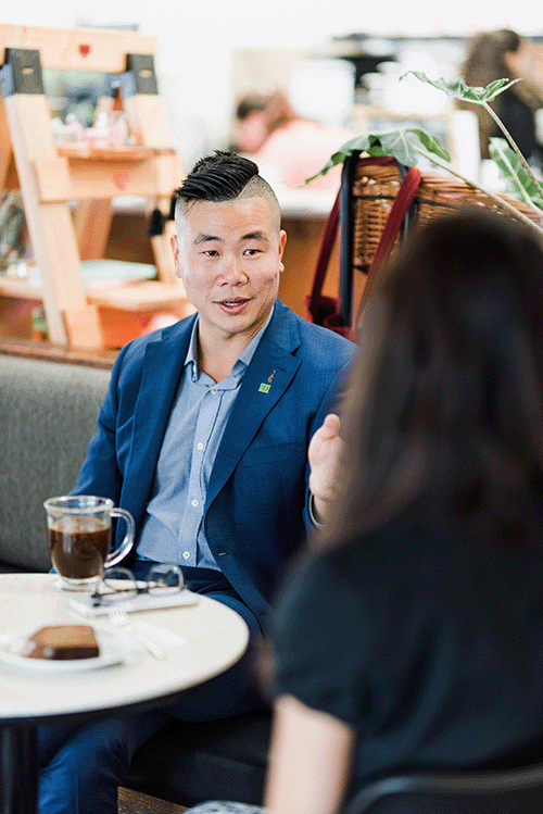 Fred Zhou sitting in a coffee shop with an interviewer. He is wearing a medium blue suit